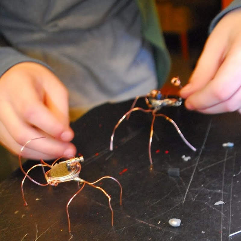 Child's hands assembling a bug bot made from electronic components and copper wire legs on a workshop table