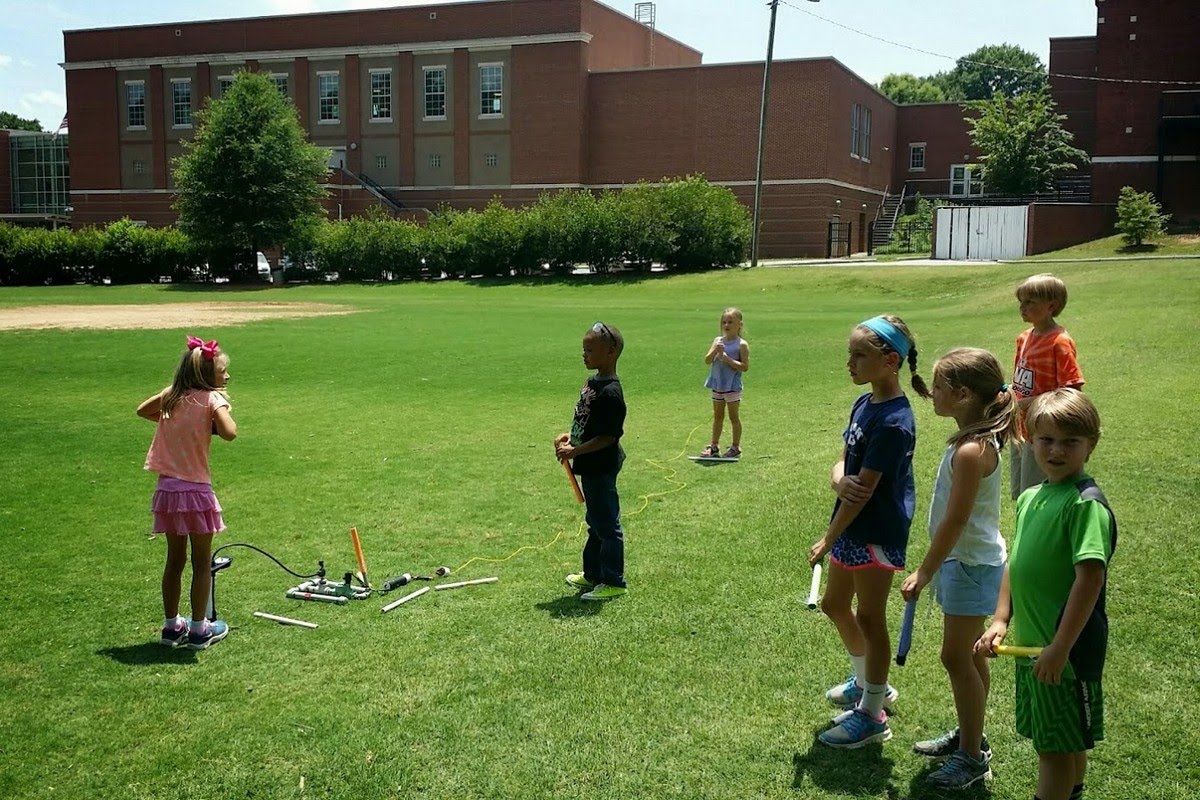 Children lined up on a school field holding their handmade rockets, waiting their turn at the compressed air launcher on a sunny summer day