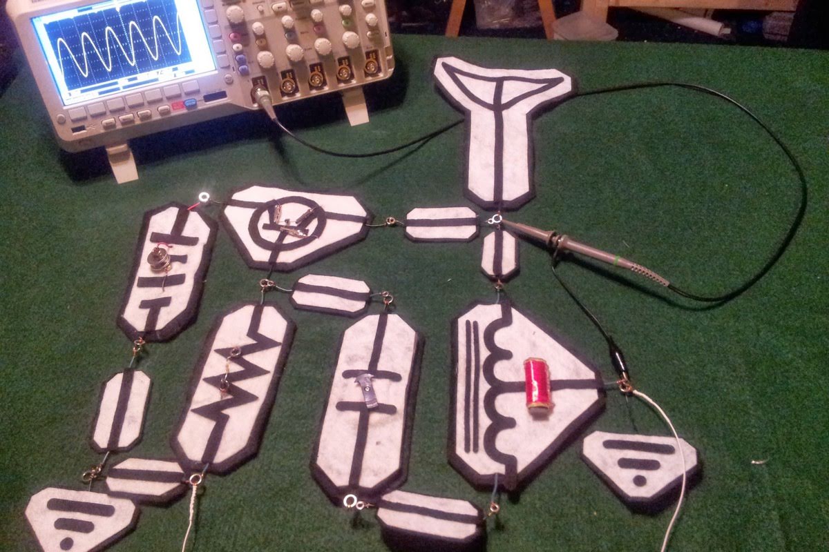Feltronics radio kit with felt circuit board pieces showing electronic symbols, connected to an oscilloscope displaying waveforms