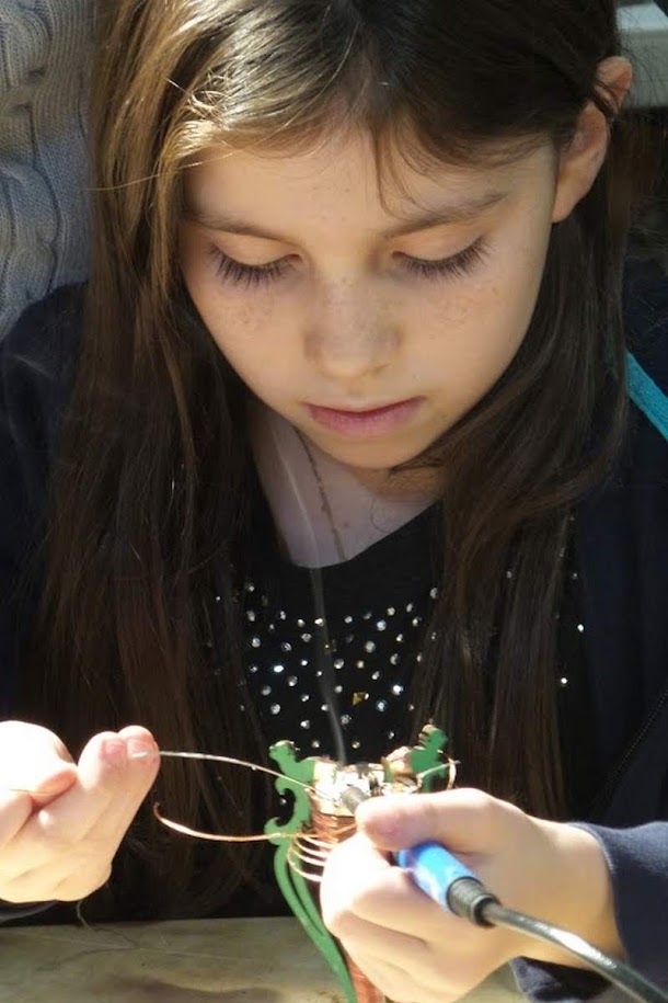 Young girl concentrating intently while soldering a mini Tesla coil circuit, learning hands-on electronics skills