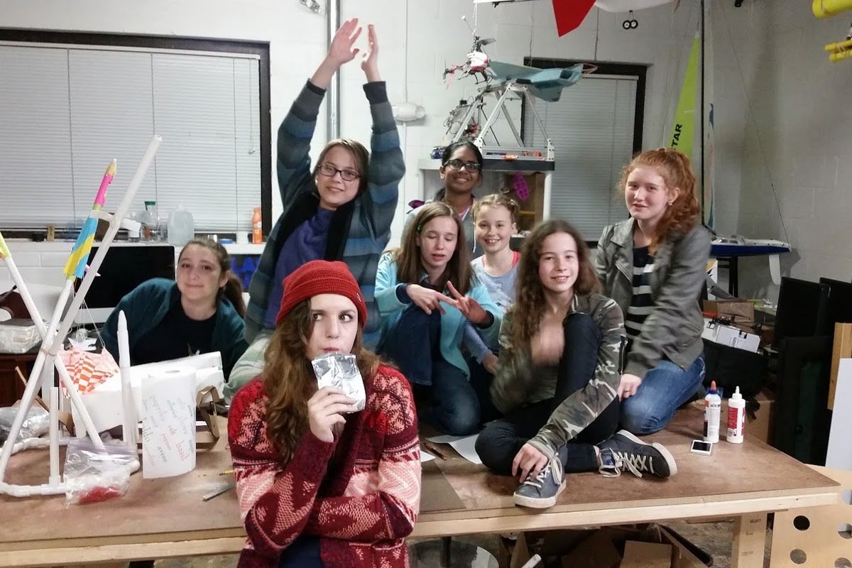 Group of seven girls in a makerspace workshop posing with their rocket and robotics projects, with a quadcopter drone visible in the background