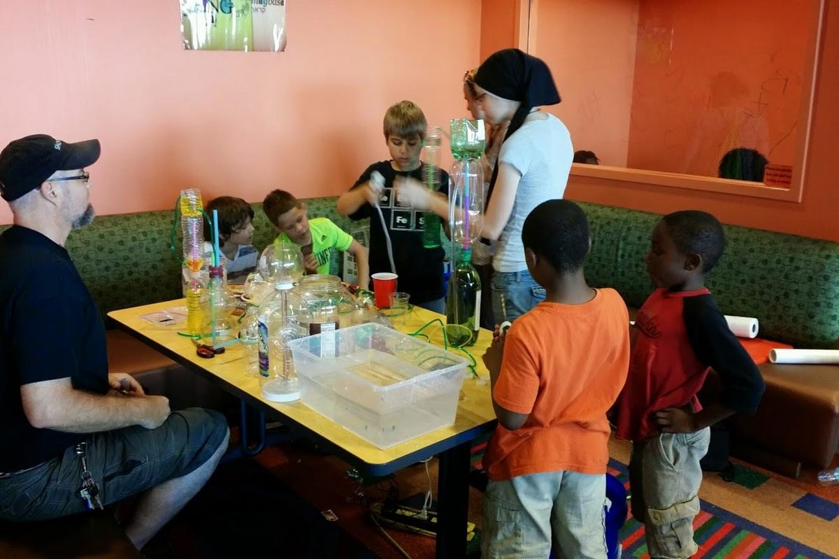 Kids gathered around a table building Heron's fountains from plastic bottles and tubes, learning about air pressure and water physics