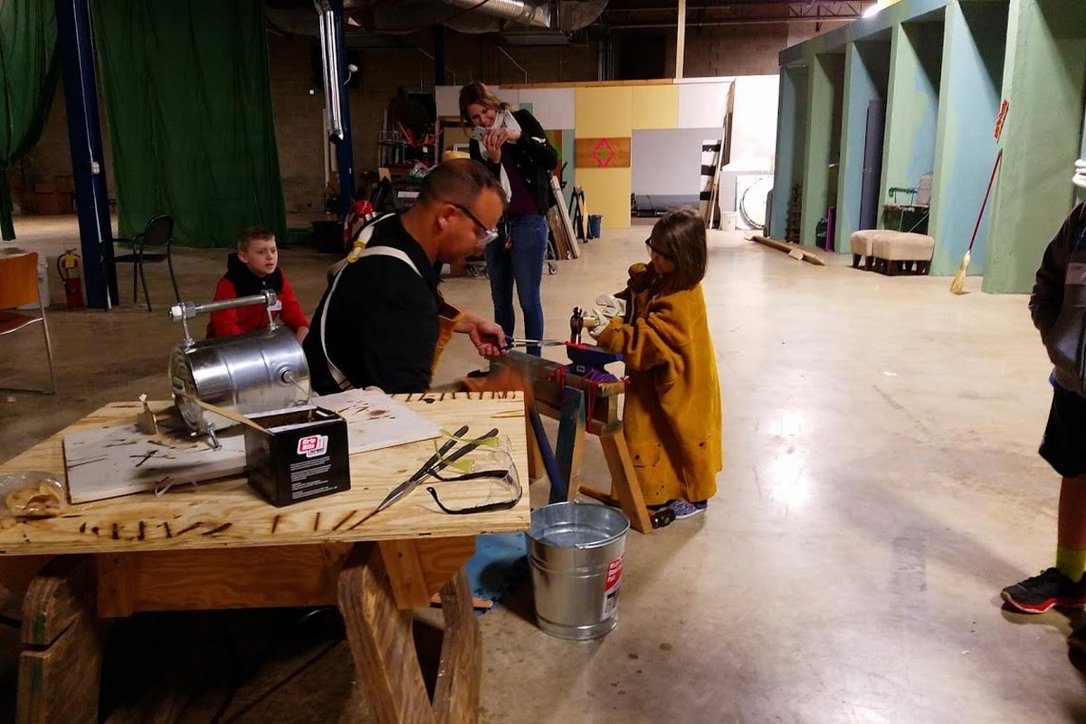 Instructor demonstrating metalworking at a forge station while a young girl in protective gear watches, with handmade mini swords displayed on the workbench