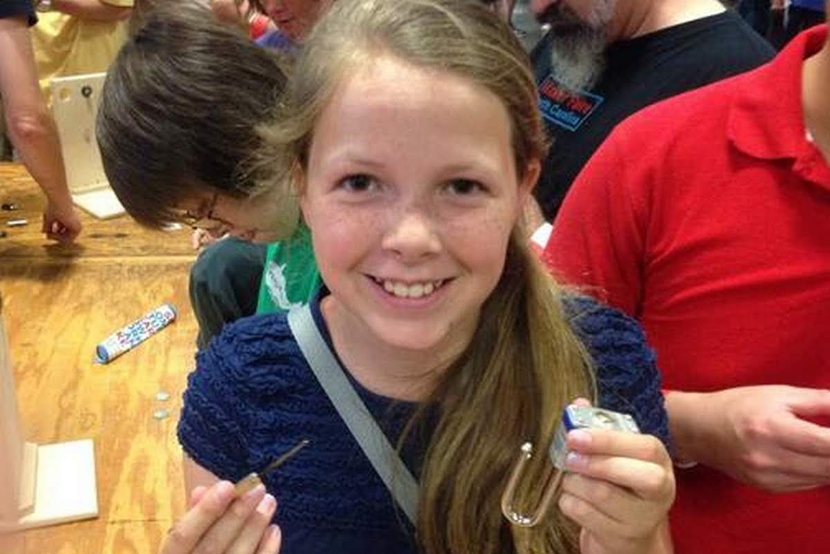 Smiling girl proudly holding up lock picks and a practice padlock she successfully picked, learning about physical security at a maker event