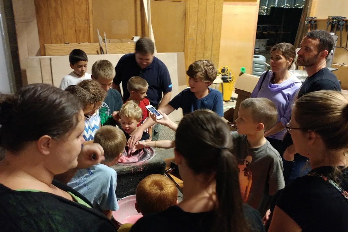 Large group of kids and parents gathered around a speaker playing non-Newtonian fluid, reaching in to touch the strange substance that acts like both liquid and solid
