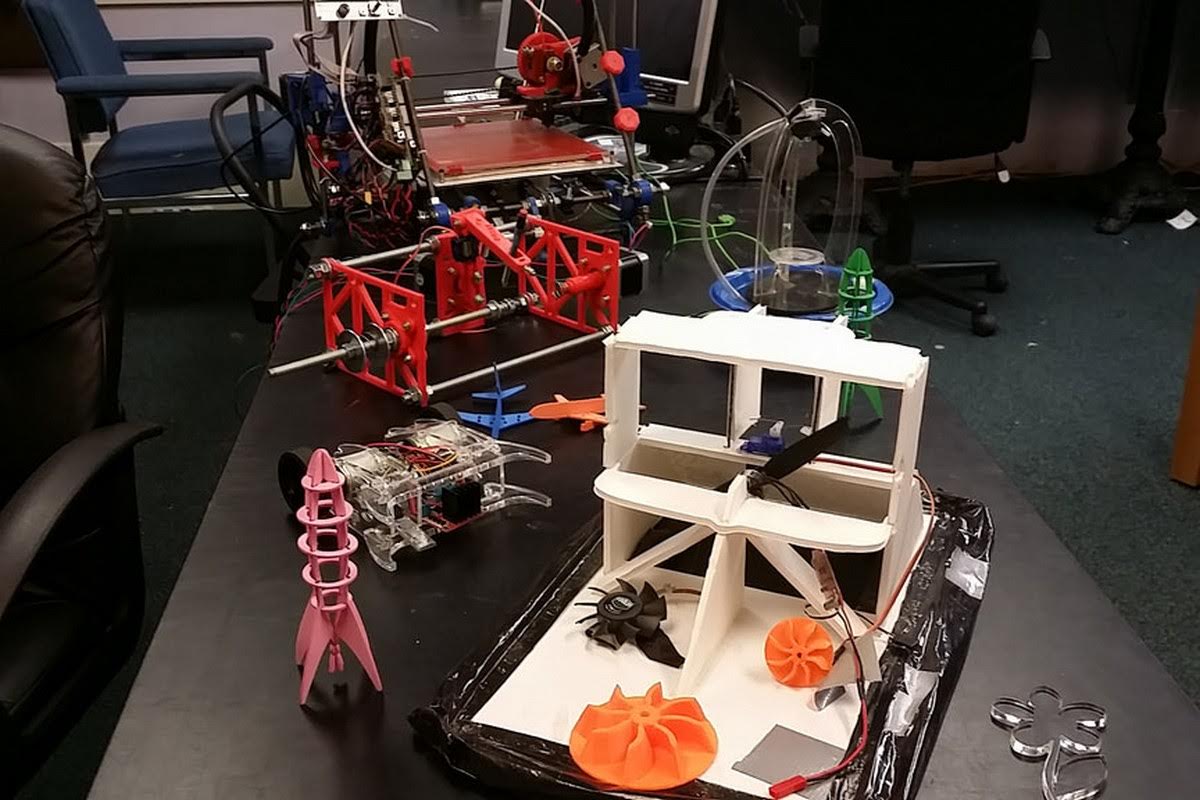 Maker workshop table displaying various student projects including RC hovercrafts with 3D printed propellers, a time fountain, 3D printed rockets, and quadcopter parts